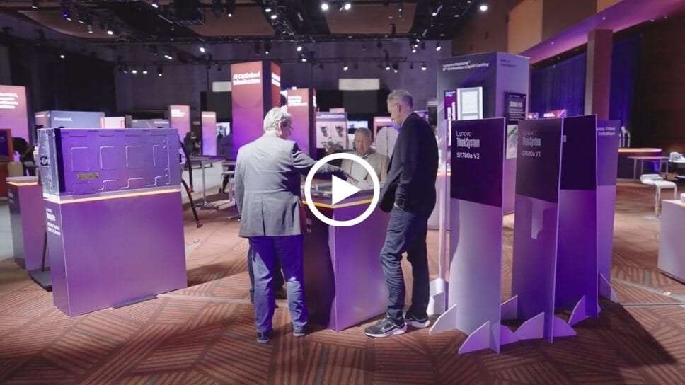 Image shows three men standing next to a desk inside of an exhibition hall. There’s a ‘play’ icon in the middle of the image.