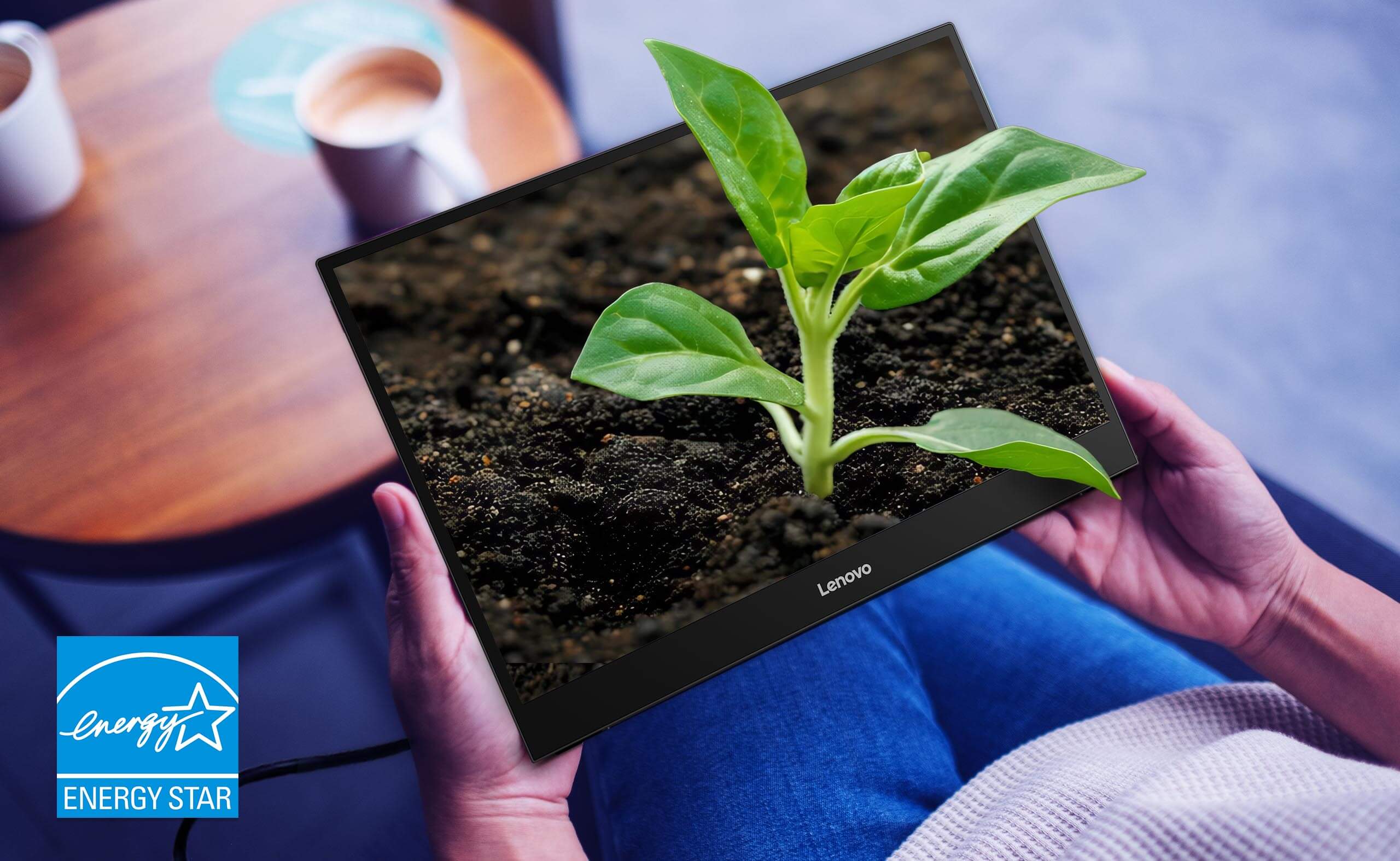 Person holding a Lenovo screen showing a vibrant green plant growing from soil. A coffee cup sits on a wooden table. Energy Star logo is visible.