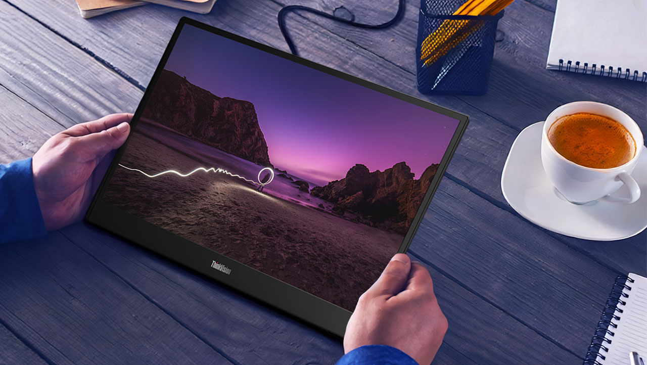 Hands holding a screen displaying a serene beach scene during sunset; a coffee cup, notepad, and pencil holder sit on a wooden table nearby.