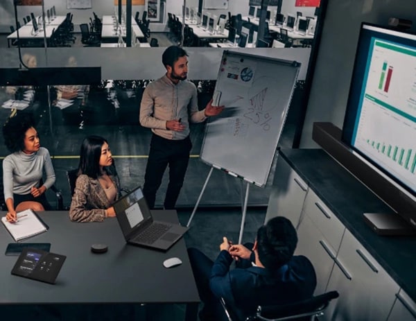 4 people in a conference room with one person standing at a white board and a bar graph displayed on TV monitor.