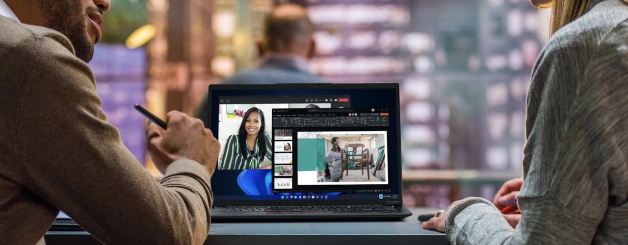 Zwei Personen an einem Tisch mit dem Lenovo ThinkPad X1 Carbon Gen 11 Notebook geöffnet zwischen ihnen und einer Videokonferenz auf dem Display.