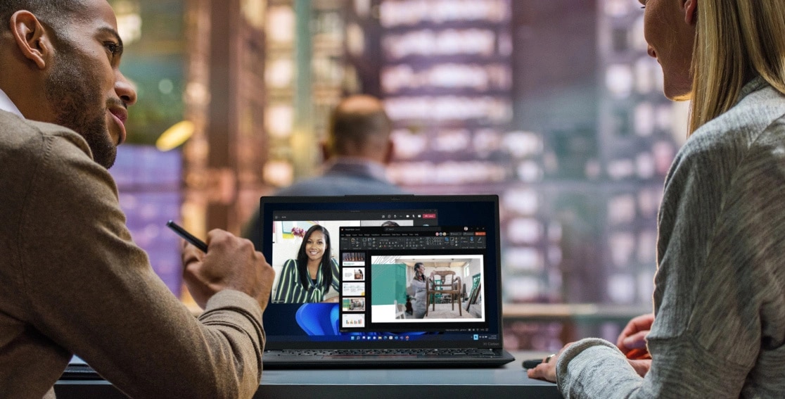 Two people at a table with the Lenovo ThinkPad X1 Carbon Gen 11 laptop open between them, with a video conference on the display.