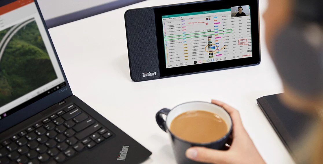 ThinkSmart View Zoom sur un bureau blanc à côté d’un portable ThinkPad avec une tasse de café dans la main.